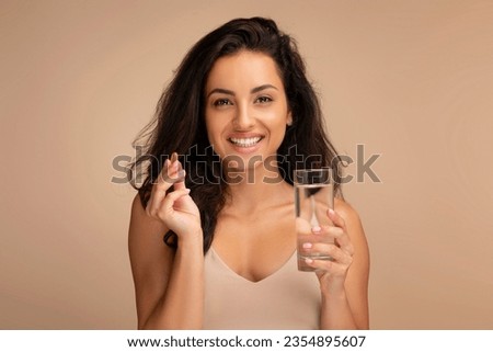 Beauty portrait of happy lovely long-haired millennial indian woman wearing beige top standing isolated over studio background, showing beauty pill capsule, supplement, holding glass of water