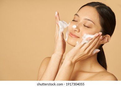 Beauty portrait of happy young woman smiling with eyes closed while applying gentle foam facial cleanser isolated over beige background