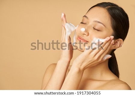 Beauty portrait of happy young woman smiling with eyes closed while applying gentle foam facial cleanser isolated over beige background