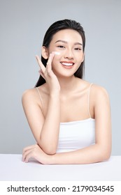 Beauty portrait image of pretty asian woman smiling and applying face cream isolated over light grey background