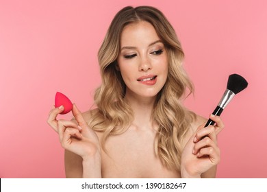 Beauty portrait of a lovely young woman with long blonde hair standing isolated over pink background, using makeup sponge