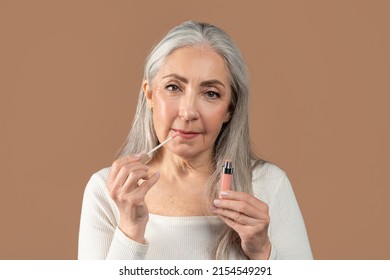 Beauty Portrait Of Senior Woman Applying Lip Gloss And Looking At Camera On Brown Studio Background. Charming Mature Lady Using Natural Decorative Cosmetics. Organic Makeup Concept