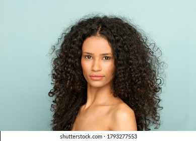Beauty portrait of a young black woman with bare shoulders and long curly hair turning to look thoughtfully at the camera with a serious expression against a blue studio background