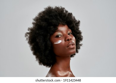 Beauty Portrait Of Young Naked African American Woman With Afro Hair Looking At Camera While Posing With Cream Applied On Her Face Isolated Over Gray Background. Skin Care Concept