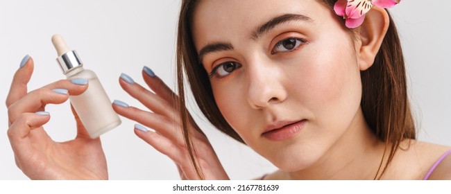 Beauty Portrait Of A Young White Red Haired Woman With Flower In Her Hair Over White Wall Background Holding Bottle Of Facial Serum Oil