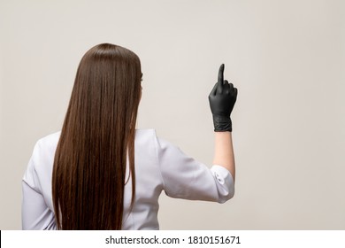 Beauty Procedure. Master Class. Brunette Woman In White Medical Overall Black Latex Gloves Pointing At Copy Space Isolated On Light Background. Professional Microblading. Rejuvenation Cosmetology.
