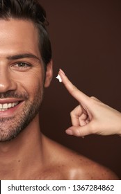 Beauty procedure and skin care. Close up half face portrait happy smiling handsome man looking at camera while female going to apply face cream on him. Isolated on brown