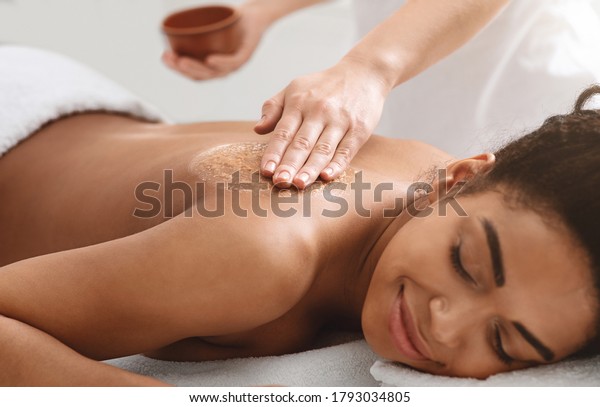 Beauty procedures at spa. Masseuse applying body\
scrub on black girl back,\
closeup