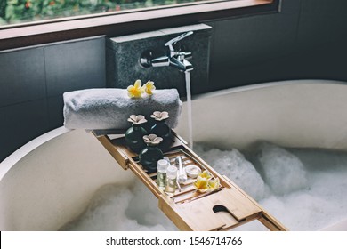 Beauty products on tray on bathtub. Bath with foam is filling with water. Relaxing in Bali luxury spa hotel.