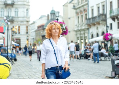 Beauty red-haired woman with curled hair in casual clothing walking on market square in old town. Confident woman in white shirt, jeans, purse walk in downtown at spring day, urban life style