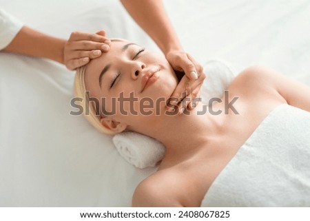 Beauty And Relaxation. Young blonde woman enjoying facial massage, lying on spa bed indoor at beauty salon, high angle shot of lady relaxing during facebuilding treatment