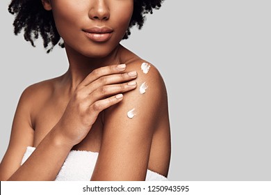 Beauty routine. Close up of young African woman applying body cream and smiling while standing against grey background
