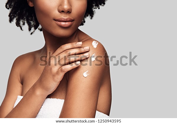 Beauty\
routine. Close up of young African woman applying body cream and\
smiling while standing against grey\
background