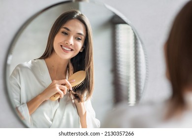 Beauty Routine. Pretty Woman Combing Her Beautiful Hair With Brush While Standing Near Mirror In Bathroom, Attractive Young Lady Looking To Her Reflection And Smiling, Selective Focus With Free Space