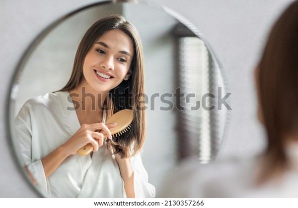 Beauty Routine. Pretty Woman Combing Her Beautiful\
Hair With Brush While Standing Near Mirror In Bathroom, Attractive\
Young Lady Looking To Her Reflection And Smiling, Selective Focus\
With Free Space