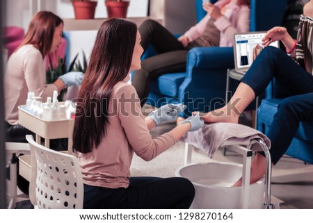 In the beauty salon. Joyful dark haired woman sitting near the client while doing the pedicure for her client