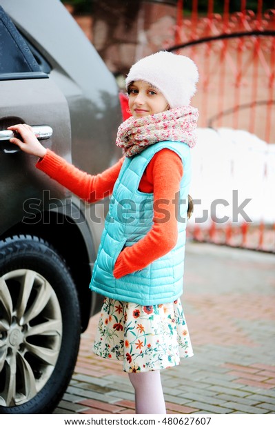 Beauty school aged kid girl in colorful outfit hat\
and warm scarf near the\
car