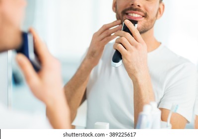 beauty, shaving, grooming and people concept - close up of young man looking to mirror and shaving beard with trimmer or electric shaver at home bathroom - Shutterstock ID 389225617