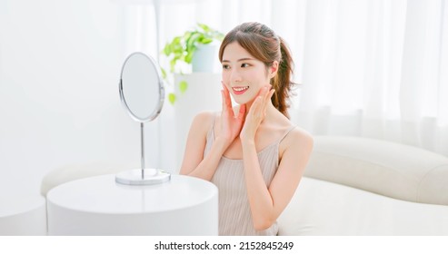 beauty skin care asian woman with brunette ponytail both hands touches her face smiling and looking into mirror