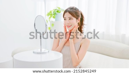 beauty skin care asian woman with brunette ponytail both hands touches her face smiling and looking into mirror
