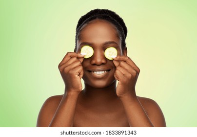 Beauty, Skin Care And Detox Concept - Beautiful African American Woman Making Eye Mask Of Cucumbers Over Lime Green Natural Background