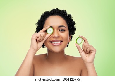 Beauty, Skin Care And Detox Concept - Beautiful African American Woman Making Eye Mask Of Cucumbers Over Lime Green Natural Background