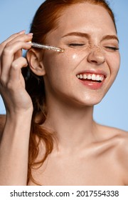 Beauty skin care. Laughing redhead girl apply c-serum with dropper on cheek, smiling white teeth. Happy woman taking care of facial skin with cosmetic products, at home cosmetology