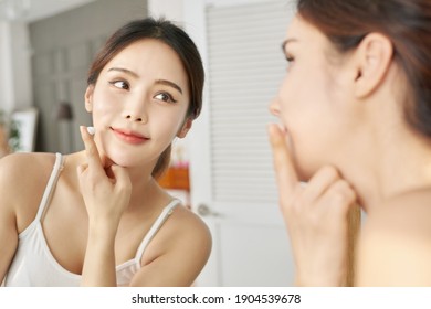 Beauty, Skin Care And People Concept - Smiling Young Woman Applying Cream To Face And Looking To Mirror At Home Bathroom