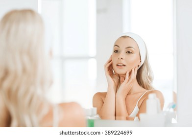 美容・スキンケア・人々のコンセプト – 顔にヘアバンドをかけた若い女性がほほ笑み、自宅のトイレで鏡を見る