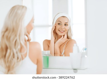 Beauty, Skin Care And People Concept - Smiling Young Woman In Hairband Touching Her Face And Looking To Mirror At Home Bathroom