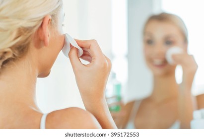 beauty, skin care and people concept - close up of smiling young woman cleaning her face with cotton disc and lotion at bathroom