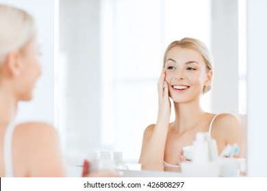 Beauty, Skin Care And People Concept - Smiling Young Woman Applying Cream To Face And Looking To Mirror At Home Bathroom