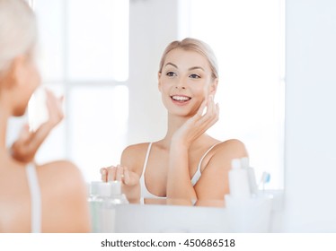 Beauty, Skin Care And People Concept - Smiling Young Woman Applying Cream To Face And Looking To Mirror At Home Bathroom