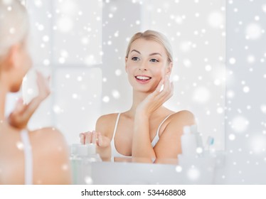 beauty, skin care and people concept - smiling young woman applying cream to face and looking to mirror at home bathroom over snow