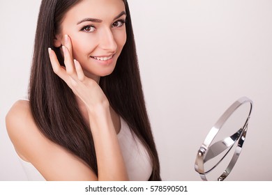 Beauty, Skin Care And People Concept - Smiling Young Woman Applying Cream To Face And Looking To Mirror At Home Bathroom.