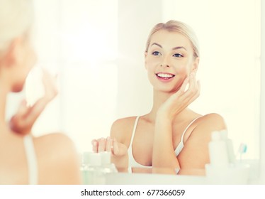 Beauty, Skin Care And People Concept - Smiling Young Woman Applying Cream To Face And Looking To Mirror At Home Bathroom