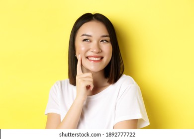 Beauty And Skincare. Close Up Of Young Asian Woman With Short Dark Hair, Healthy Glowing Skin, Smiling And Touching Dimples On Cheeks, Standing Over Yellow Background