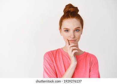 Beauty and skincare. Close-up of cute teen girl with freckles and red hair combedin messy bun, smiling thoughtful at camera, standing over white background.