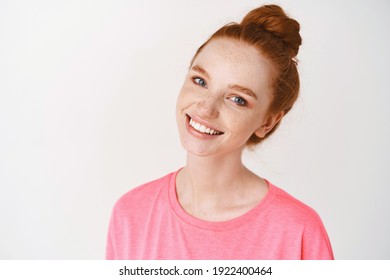 Schönheit und Hautpflege. Nahaufnahme einer jungen Rotkopffrau mit Sommersprossen und blauen Augen, die sauber berührend wirken, keine Schminkhaut und Lächeln, die auf rosafarbenem T-Shirt auf weißem Hintergrund stehen.