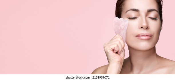 Beauty And Skincare Concept With A Beautiful Woman Doing Skin Care Routine With Gua Sha Tool. Middle Aged Female Using Guasha. Massage For Facial Lifting. Pink Background Banner