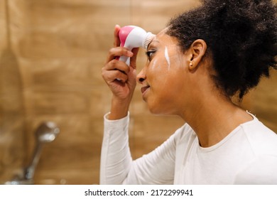 Beauty And Skincare Concept. Young African Woman Using Cleansing Foam For Washing Her Face With Silicone Brush In Bathroom Closeup