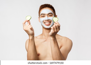 Beauty, skincare and spa concept. Happy handsome asian man with naked torso, taking care of skin, self-distancing enjoying and relaxing leisure, apply face mask and put cucumber on eyes
