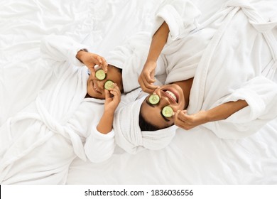 Beauty Spa Day. Cheerful Black Mom And Daughter In Bathrobes Lying With Cucumber Slices On Eyes, Doing Face Mask Treatment, Wearing Towel On Head, Having Fun Together At Home, Top View With Copy Space - Powered by Shutterstock