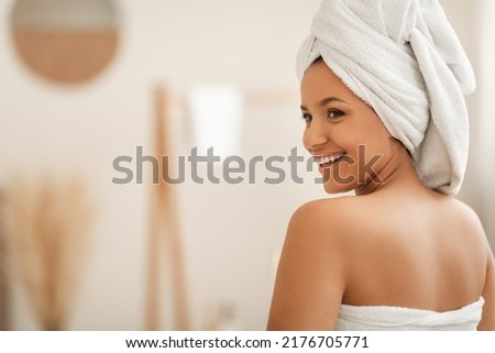 Beauty And Spa. Happy Woman Posing Wrapped In White Towel Smiling Looking Aside At Free Space Sitting Back To Camera Enjoying Bodycare Routine After Bathing In Modern Bathroom Indoors