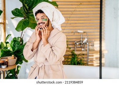 Beauty, spa and leisure concept. Funny asian man with green eyes patches in bathrobe and bath towel over head sitting on bathtub in cozy bathroom.