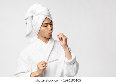 Beauty, spa and leisure concept. Portrait of handsome young man have day-off, enjoying quarantine, wear bathrobe and bath towel on head, polishing nails, standing white background