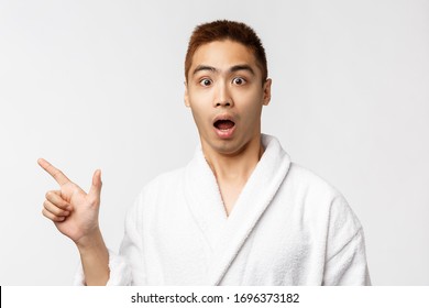 Beauty, spa and leisure concept. Portrait of amazed and startled asian man in bathrobe, open mouth intrigued and interested, pointing finger left, showing way, white background