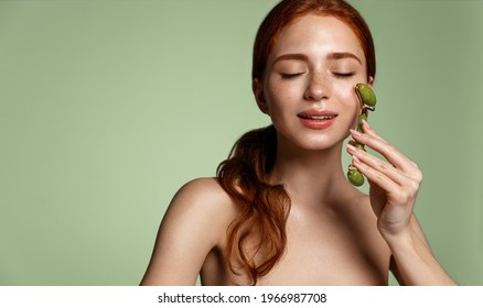 Beauty spa. Redhead woman massage facial skin with natural jade roller. Girl pleasure from using lifting tool on her face, massaging muscles to reduce tension, rubbing in c-serum skincare treatment