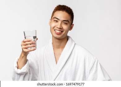 Beauty, Spa And Technology Concept. Cheers To Healthy Lifestyle. Portrait Of Cheerful Asian Man In Bathrobe Raising Glass Of Water And Smiling, Wear Bathrobe, Wake Up And Drink, White Background