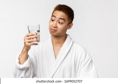 Beauty, Spa And Technology Concept. Not Bad Taste. Portrait Of Pleased Asian Man In Bathrobe Sipping Water From Glass And Look Satisfied Camera, Standing White Background